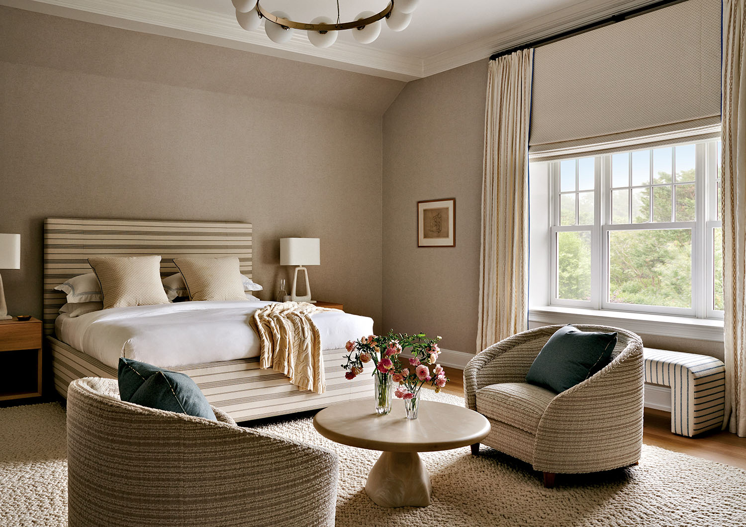 A neutral-toned bedroom with a striped bed, two armchairs, a round coffee table with flowers, large window with bench, and minimalist decor.