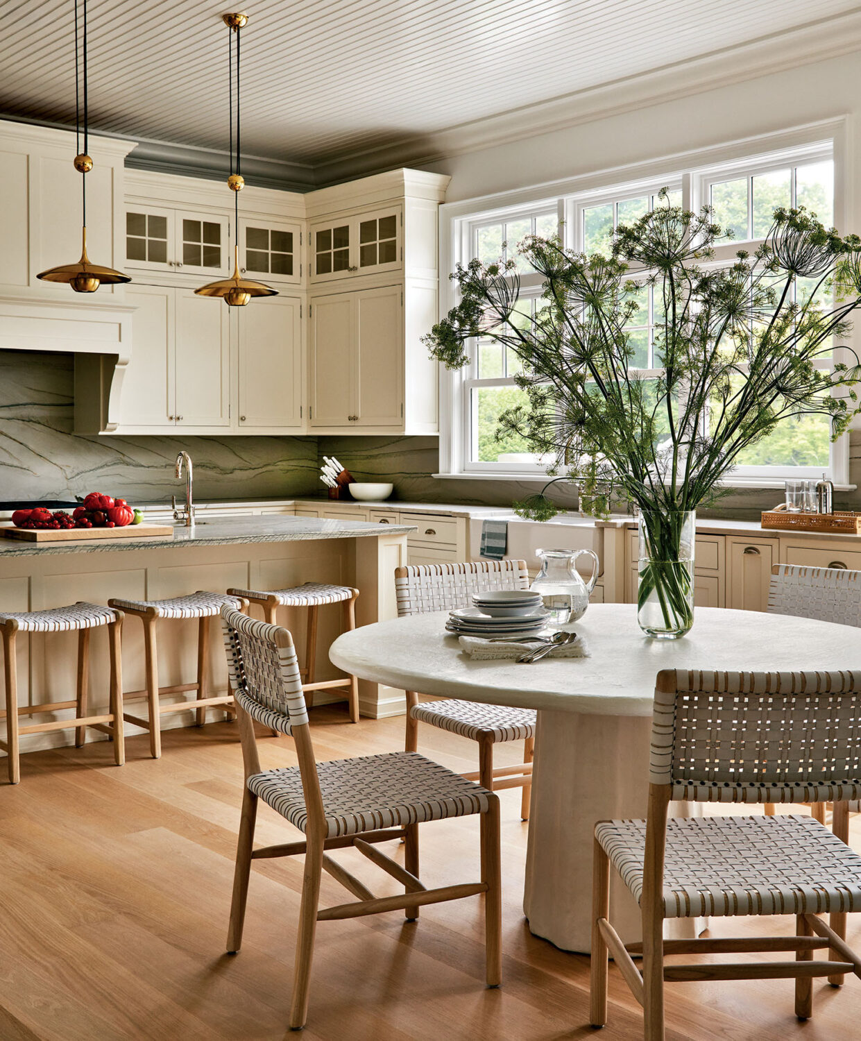 Bright kitchen with cream cabinets, marble backsplash, wooden floors, round dining table with four chairs, and a vase of tall green flowers on the table near large windows.