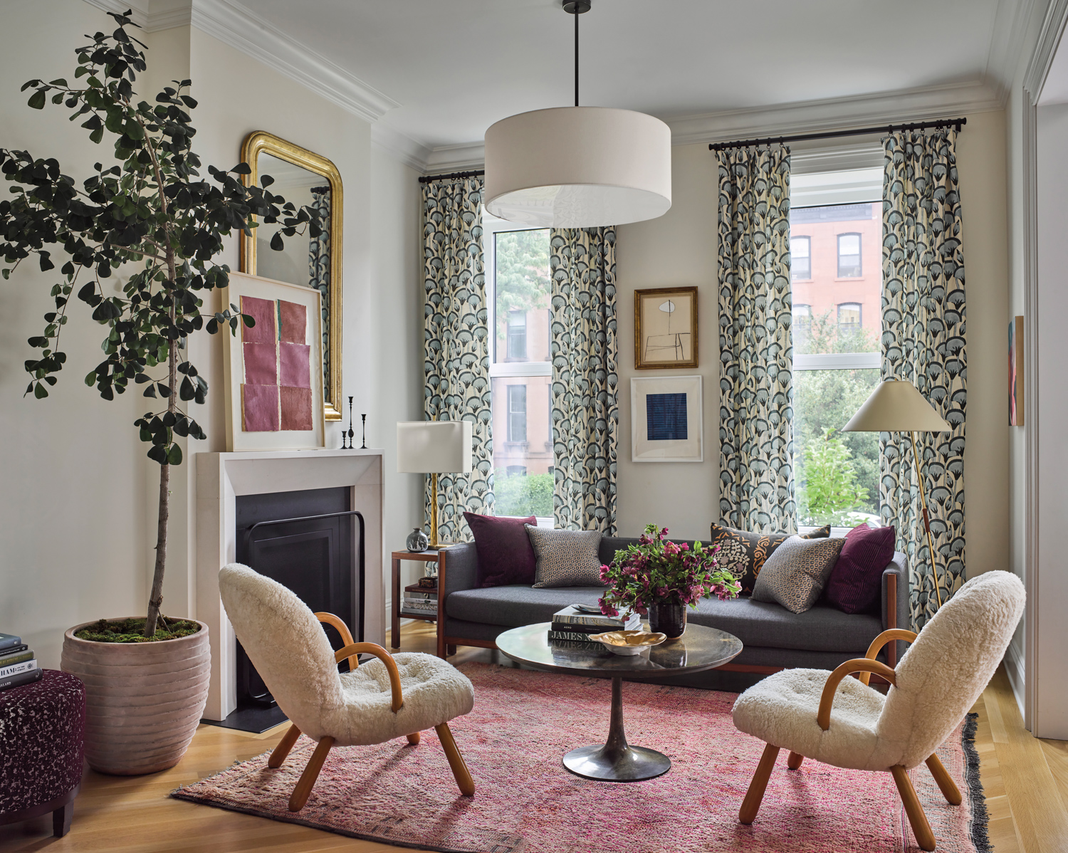 Cozy living room with a gray sofa, patterned curtains, and two plush chairs around a round coffee table. Soft lighting and art create a welcoming ambiance.