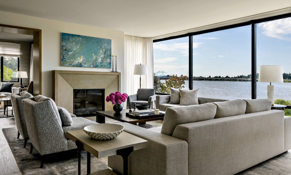 Modern living room with light gray sofas, armchairs, a fireplace, large windows overlooking water, a coffee table with decor, and a blue abstract painting above the mantel.