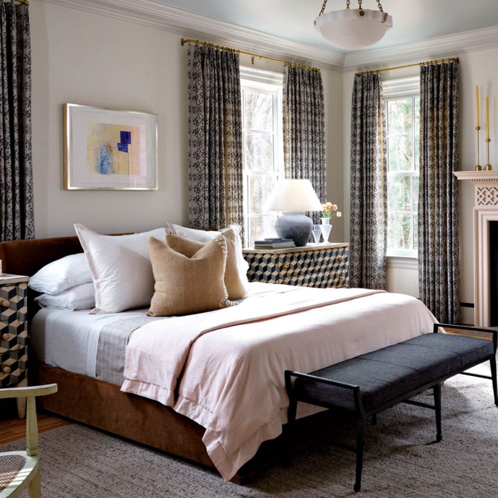 A neatly made bed with neutral bedding, two patterned nightstands, a bench at the foot, tall windows with patterned curtains, and a fireplace in a bright, stylish bedroom.