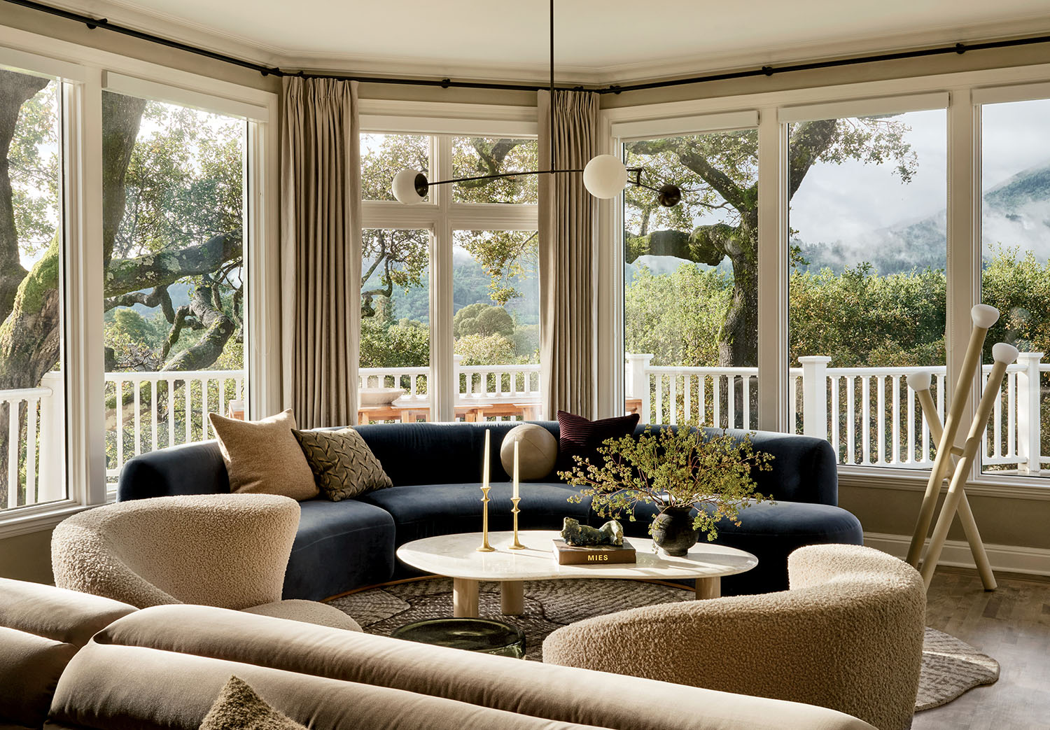A cozy living room with a curved, navy-blue sofa and curved beige chairs around a round coffee table. Large windows offer a view of trees and misty mountains.