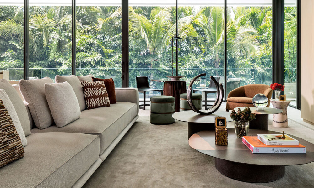 Modern living room with beige sofa, round coffee tables, armchair, and floor-to-ceiling windows overlooking lush tropical greenery. Books and decorative items are on the tables.