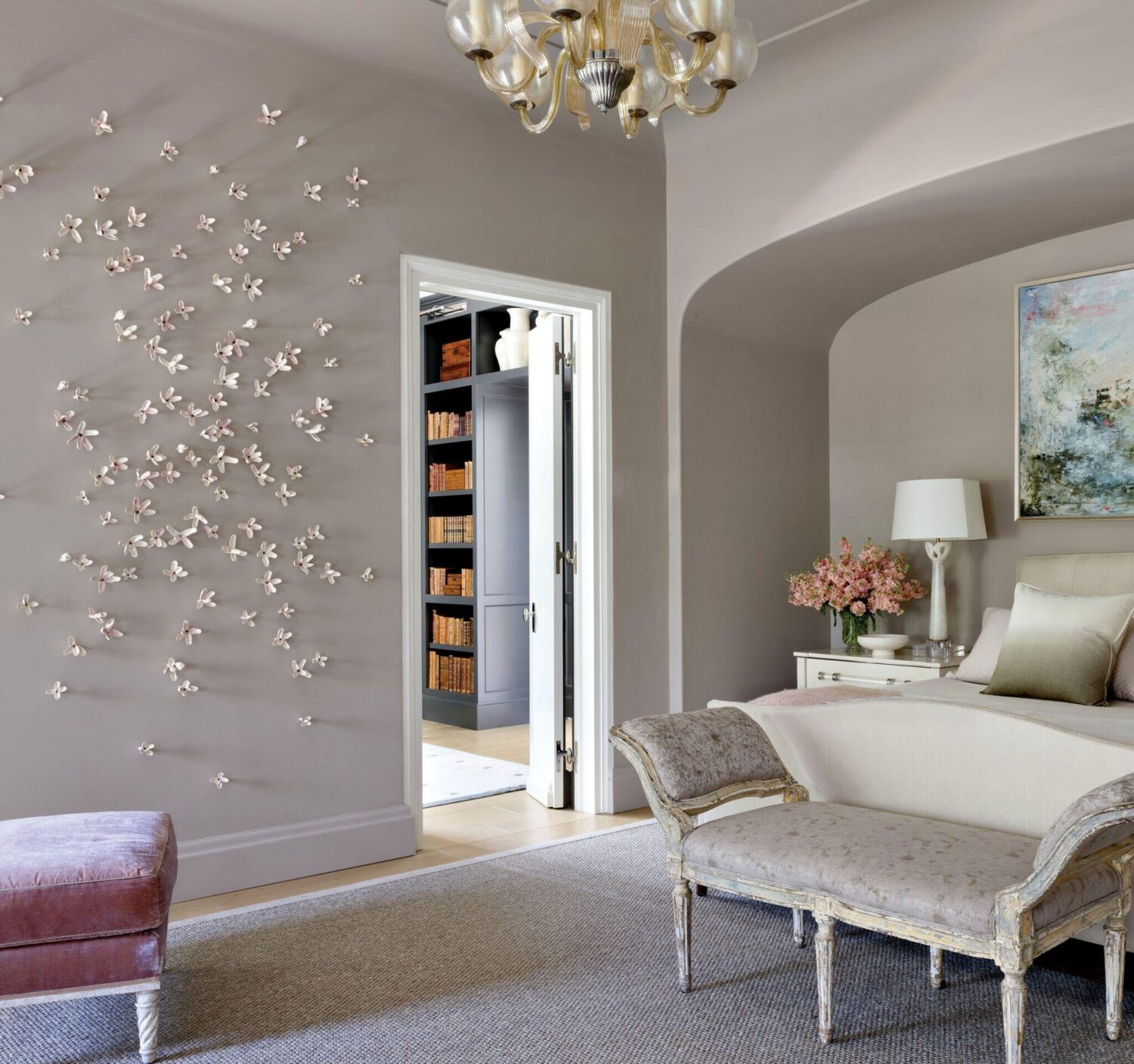 Elegant bedroom with light gray walls, decorative butterfly accents, vintage-style furniture, a chandelier, and an open door revealing a bookshelf in the next room.