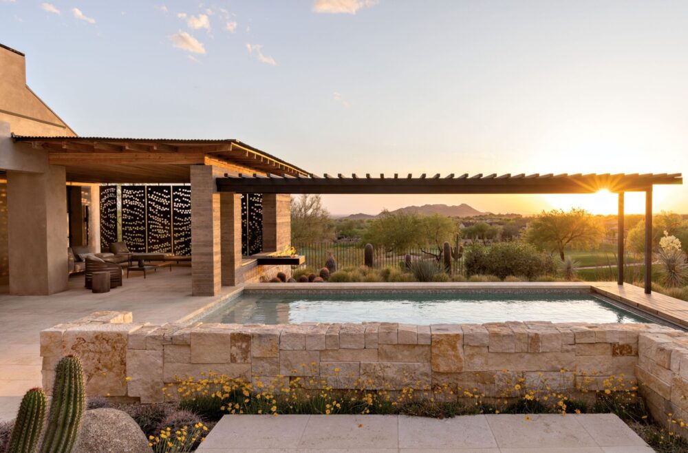 Modern backyard with a rectangular stone pool, covered patio, lounge chairs, and desert landscaping at sunset, overlooking distant mountains.