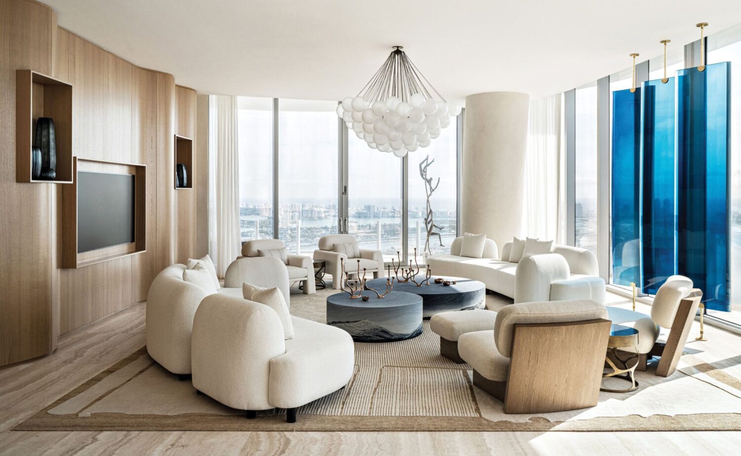 Modern living room with white curved sofas, round coffee tables, wood paneling, large windows with city view, and a sculptural chandelier. Blue glass panels hang near floor-to-ceiling windows.