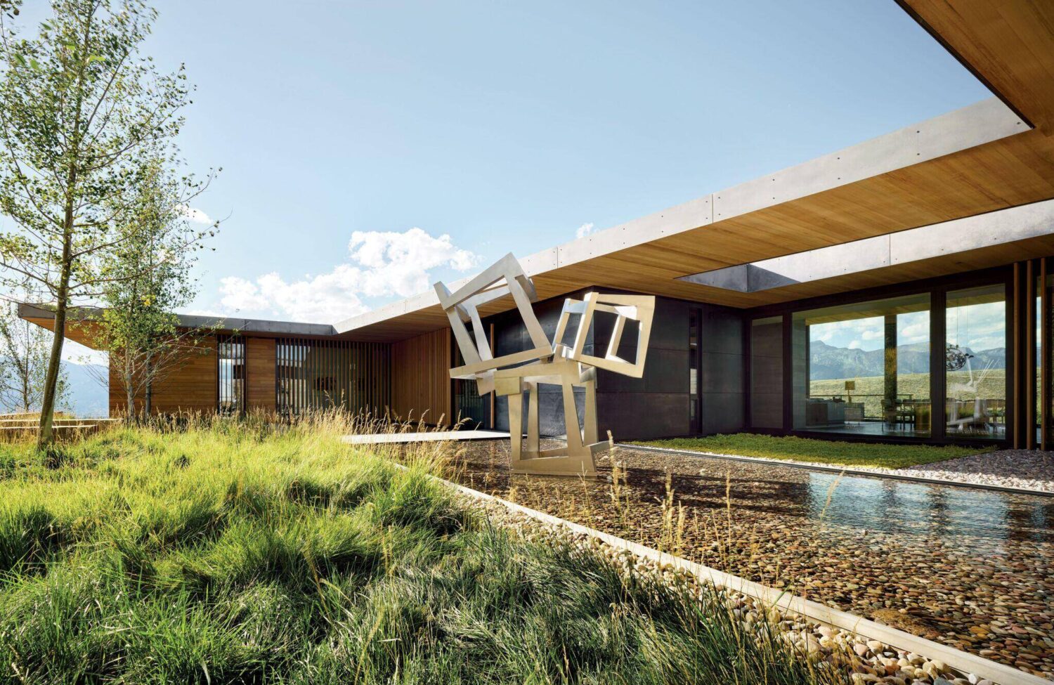 Modern house with large windows, wooden and concrete elements, grassy landscaping, and a geometric metal sculpture over a shallow water feature.