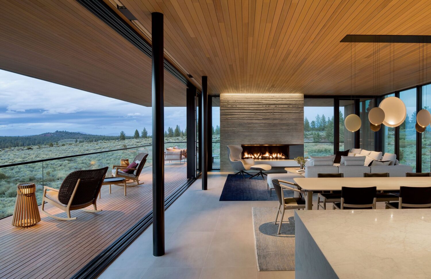 Modern open-concept living space with large windows, minimalist furniture, a fireplace, and a spacious outdoor deck overlooking a natural landscape.
