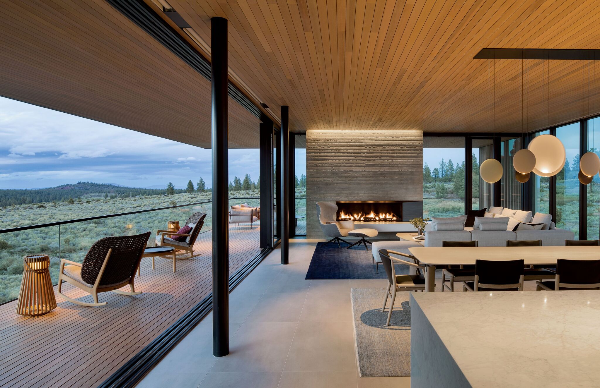 Modern open-concept living space with large windows, minimalist furniture, a fireplace, and a spacious outdoor deck overlooking a natural landscape.