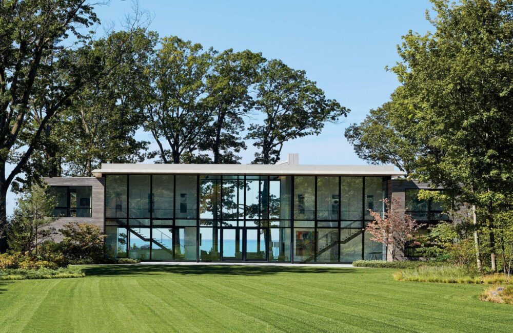 Modern two-story house with large glass windows, surrounded by trees and a well-manicured lawn, with a view of water visible through the building.