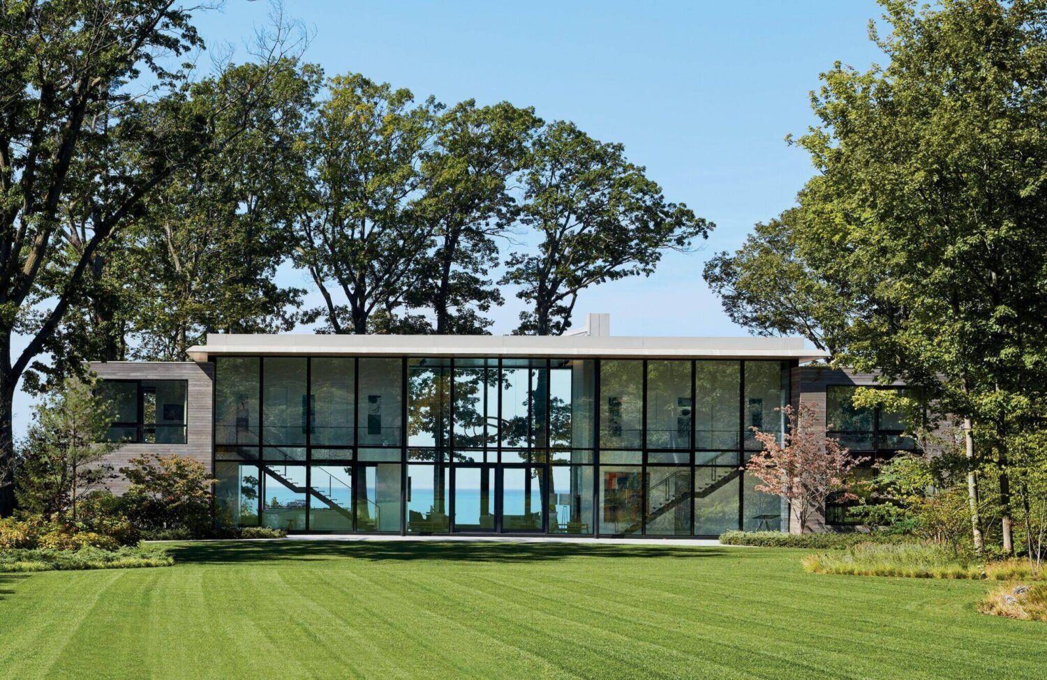 Modern two-story house with large glass windows, surrounded by trees and a well-manicured lawn, with a view of water visible through the building.