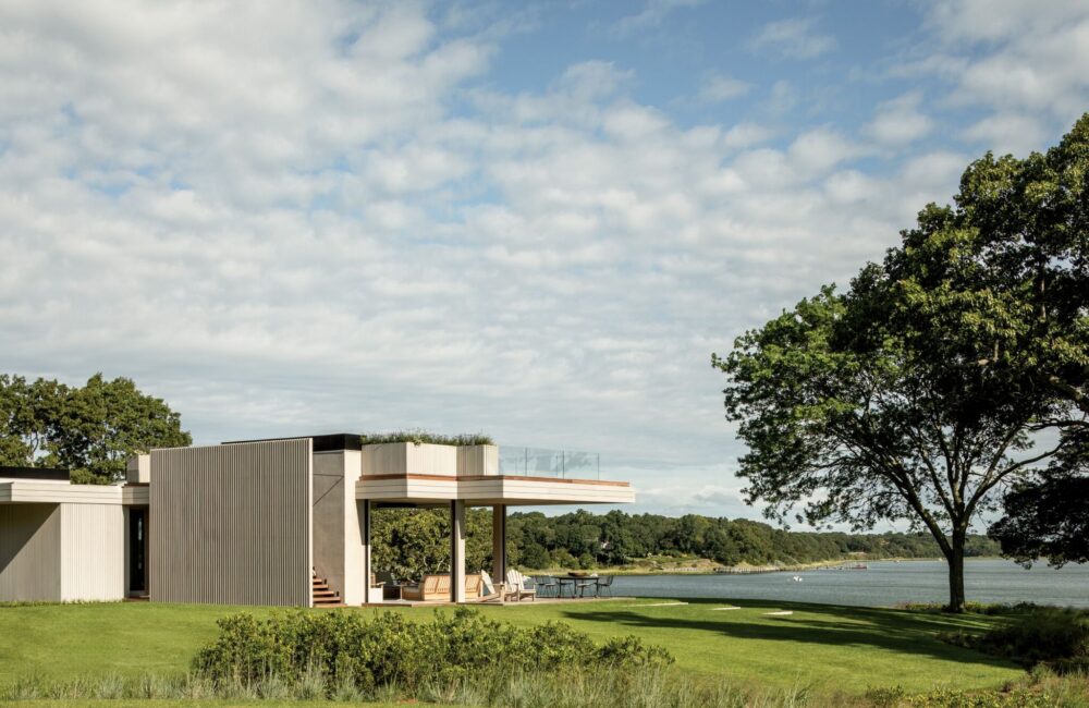 Modern house with large windows and a flat roof overlooks a grassy lawn and a body of water, surrounded by trees under a partly cloudy sky.