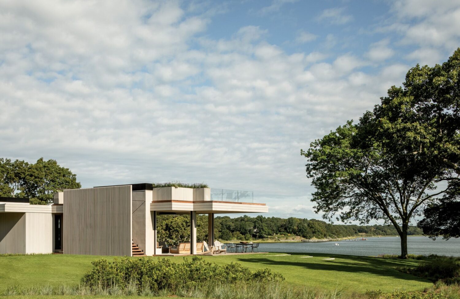 Modern house with large windows and a flat roof overlooks a grassy lawn and a body of water, surrounded by trees under a partly cloudy sky.
