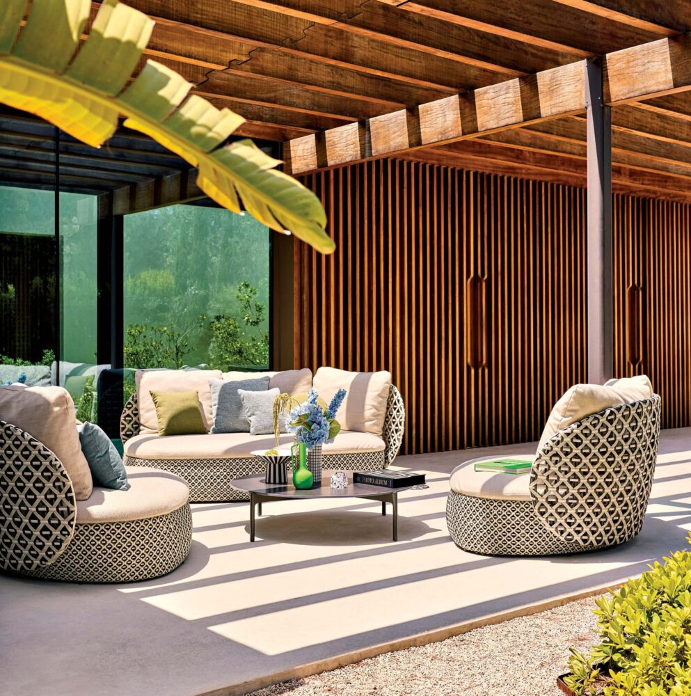 Modern outdoor patio with patterned cushioned seating, a low table with decor items, and a wooden pergola overhead, next to a building with vertical wooden slats.