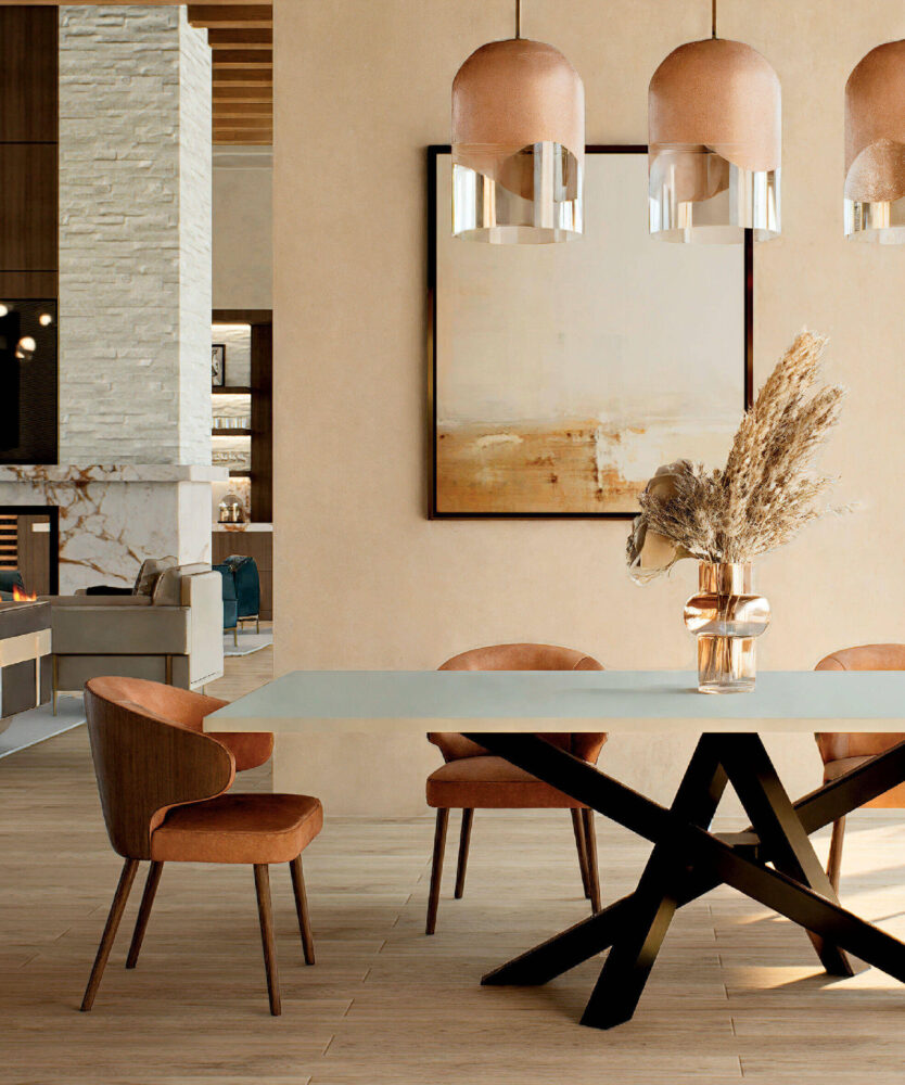 Modern dining area with a rectangular table, four upholstered chairs, three pendant lights, a vase with dried grasses, and abstract wall art. Fireplace and lounge area visible in background.