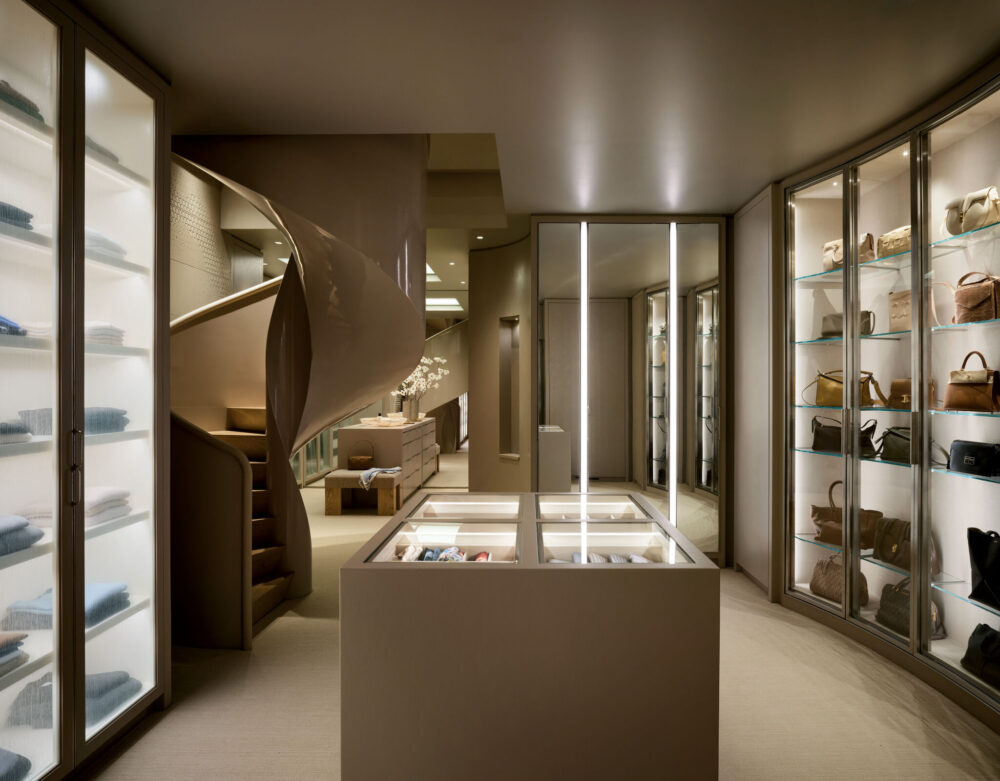 Modern boutique interior with illuminated display cases, shelves holding folded clothes and handbags, and a spiral staircase in the background.