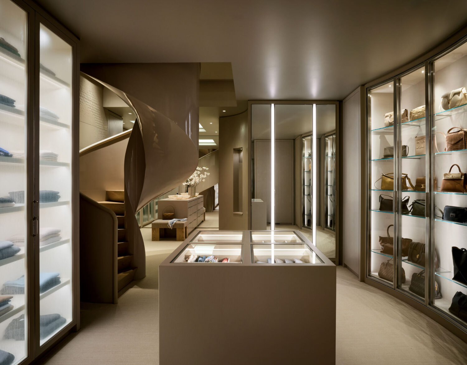 Modern boutique interior with illuminated display cases, shelves holding folded clothes and handbags, and a spiral staircase in the background.