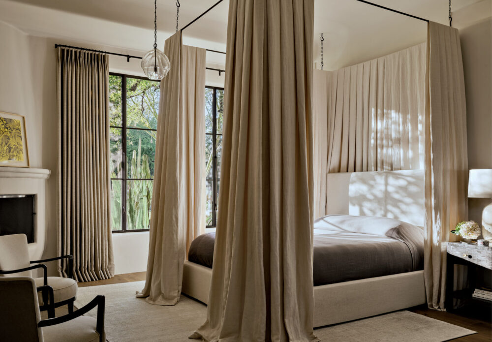 A modern bedroom with tall windows, beige curtains, a four-poster bed, upholstered chairs, a side table with a lamp, and natural light streaming in.