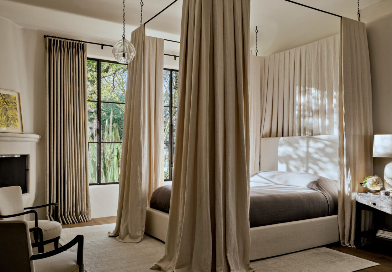 A modern bedroom with tall windows, beige curtains, a four-poster bed, upholstered chairs, a side table with a lamp, and natural light streaming in.