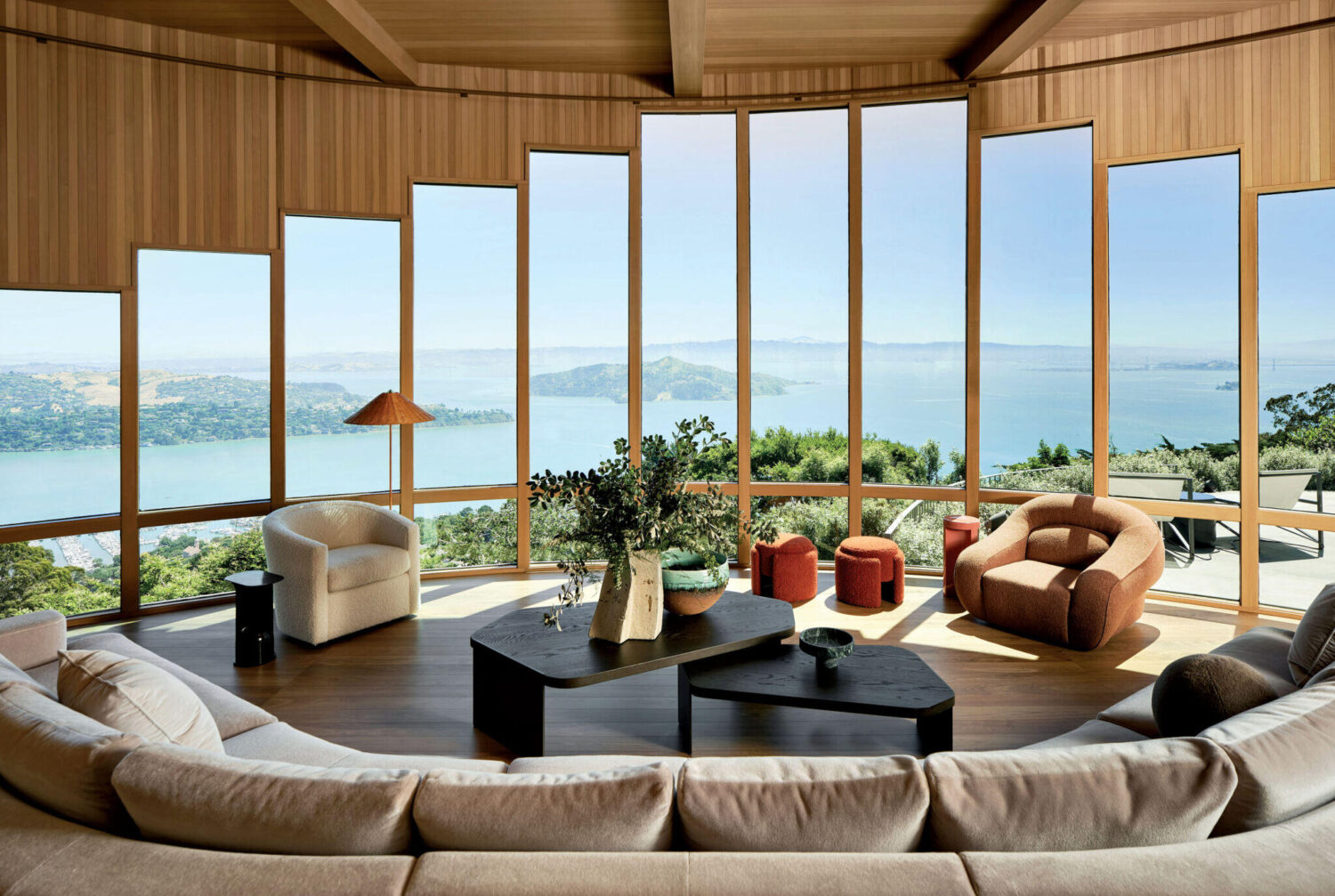 Spacious living room with curved beige sofa, modern chairs, large wooden coffee table, and floor-to-ceiling windows offering a panoramic view of a bay and distant hills.