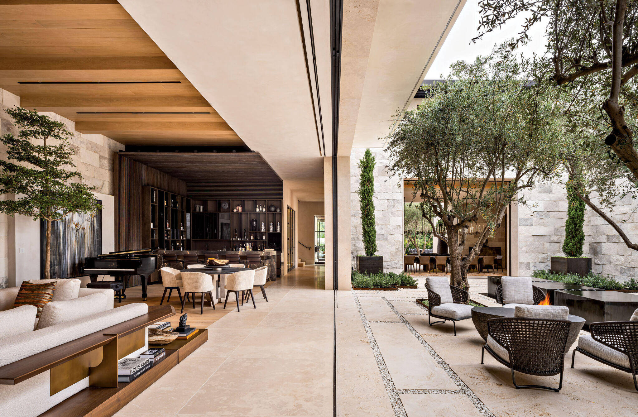 Modern open-concept living room with light stone floors, a dining area, a grand piano, and an adjoining outdoor patio with seating, trees, and a fire pit separated by glass walls.