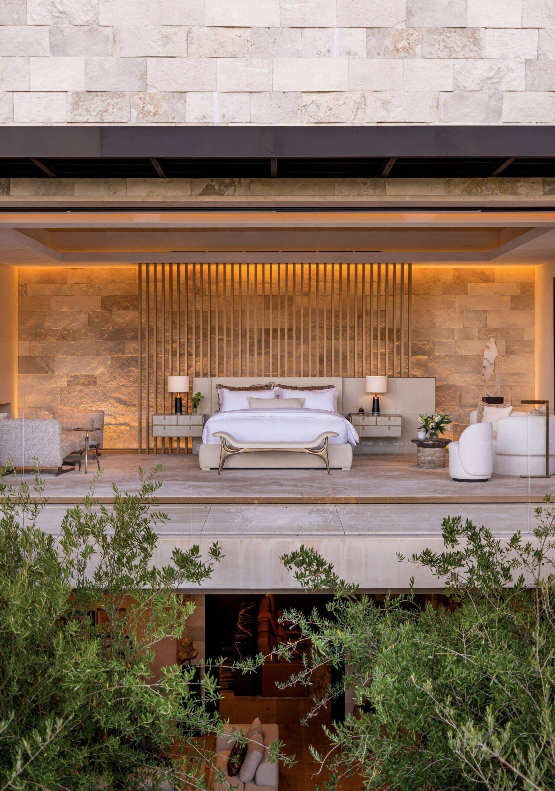 A modern open-air bedroom with a large bed, two side tables with lamps, sofas, and stone walls, overlooking greenery in the foreground.