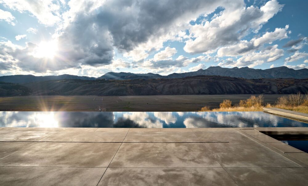 A concrete patio overlooks a reflection pool with mountains and a partly cloudy sky in the background at sunset.