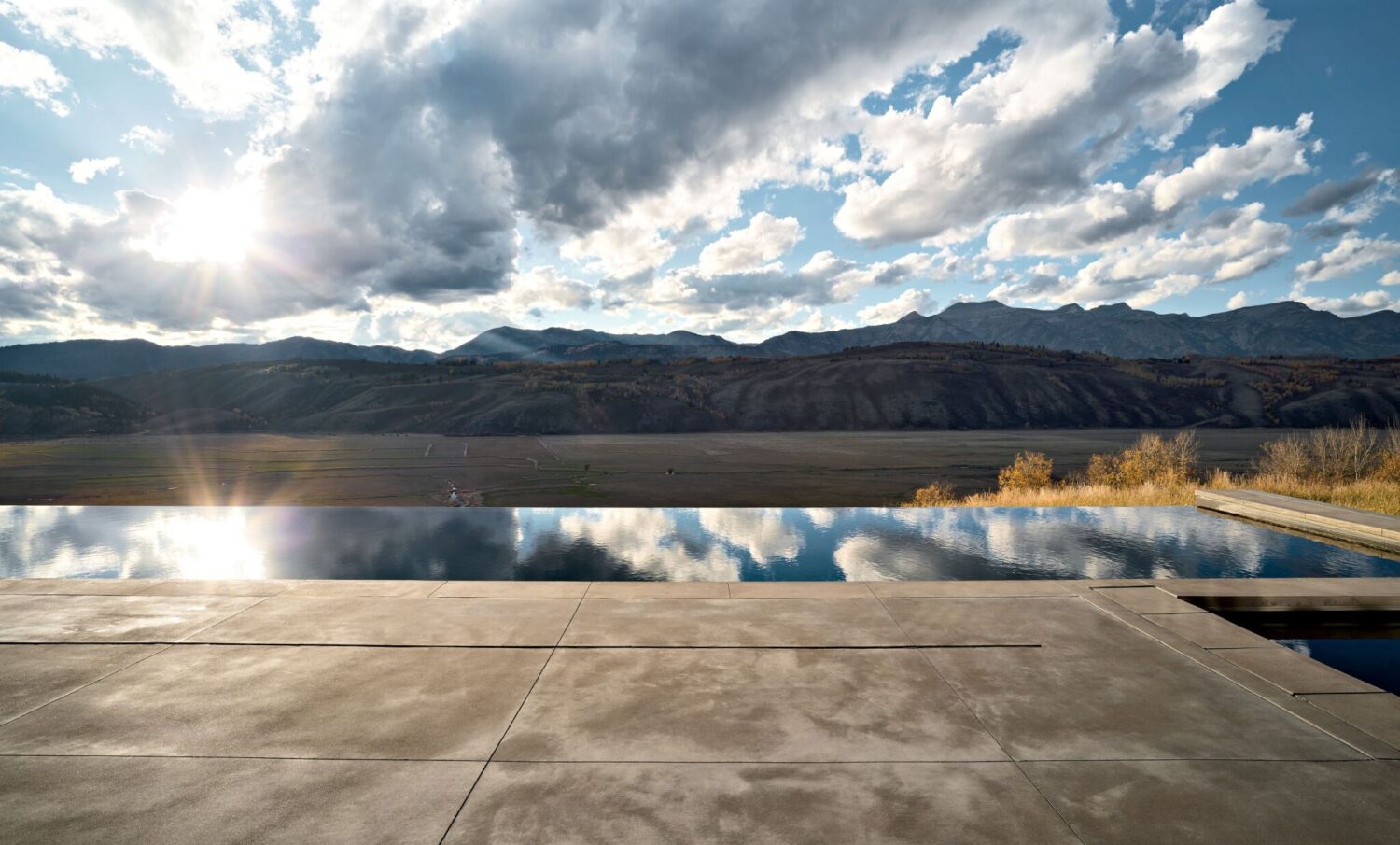 A concrete patio overlooks a reflection pool with mountains and a partly cloudy sky in the background at sunset.