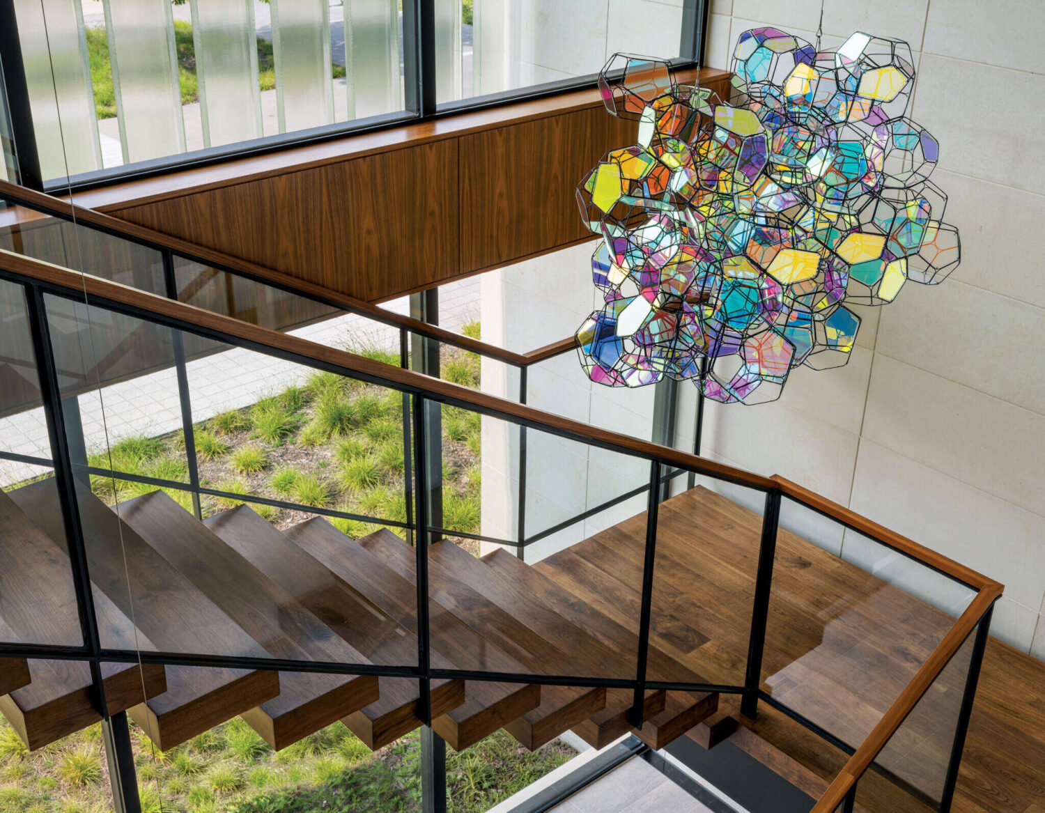 A geometric, multicolored glass sculpture hangs above a wooden staircase with black railing in a modern building with large windows.