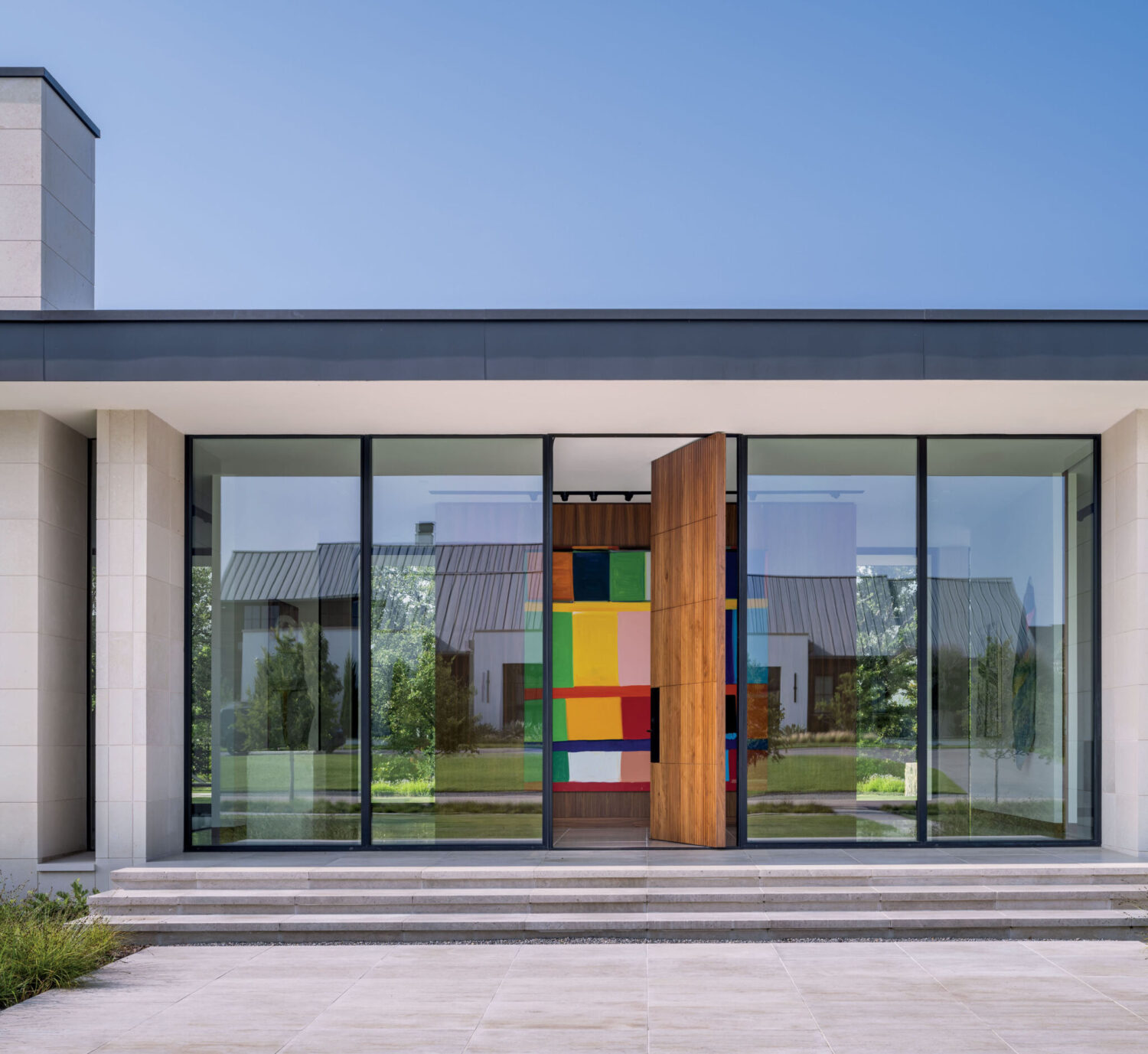 Modern building entrance with large glass panels, a pivoting wooden door, and a colorful geometric artwork visible inside; reflection of houses and trees on the glass.