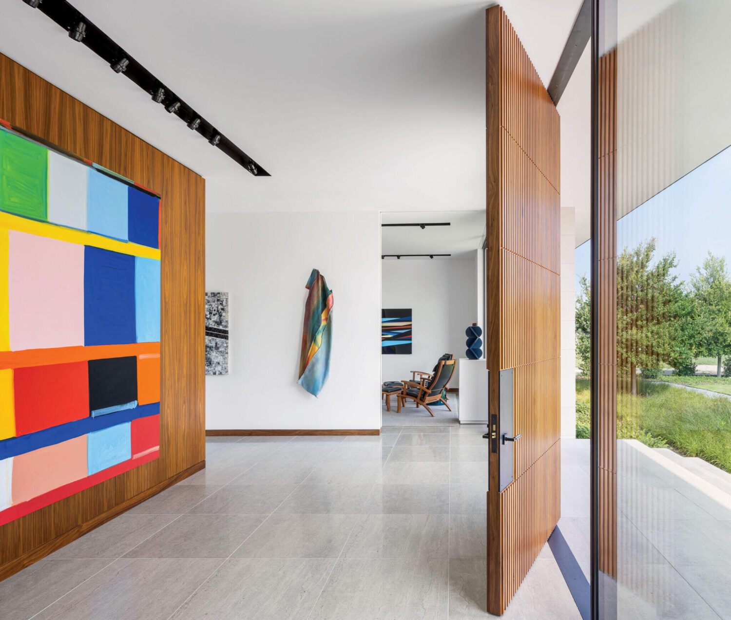 Modern entryway with large wooden door, abstract colorful wall art, tiled floor, and view into a seating area with contemporary decor and large window.