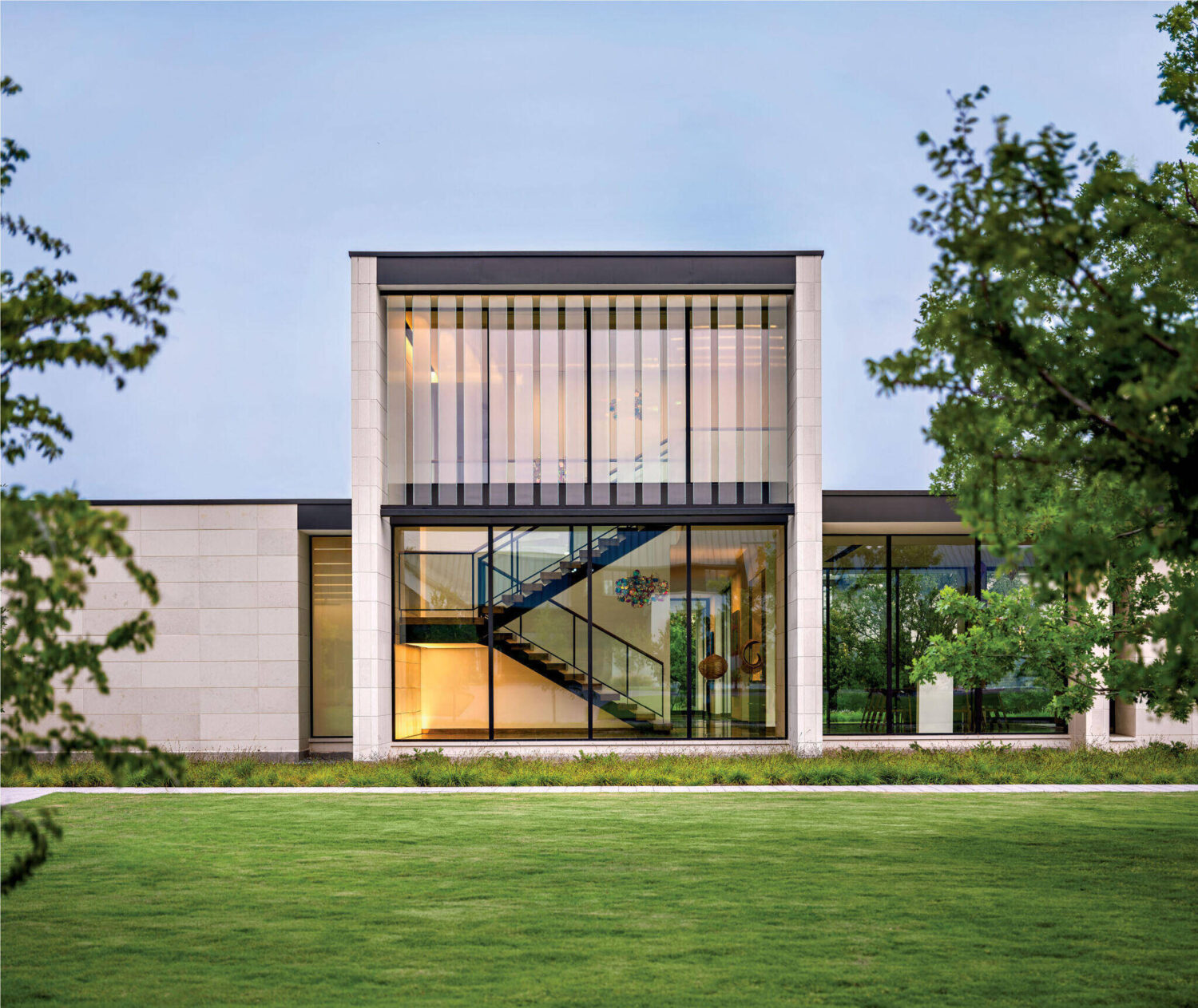 Modern two-story building with large glass windows, visible staircase, and minimalist design, surrounded by green lawn and trees.