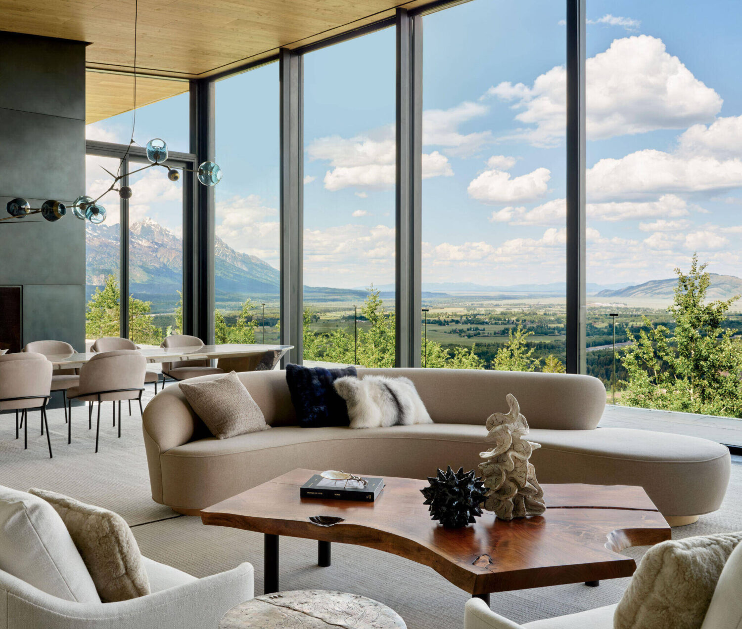 Modern living room with a curved beige sofa, artistic wooden coffee table, and large floor-to-ceiling windows offering panoramic views of mountains and greenery.