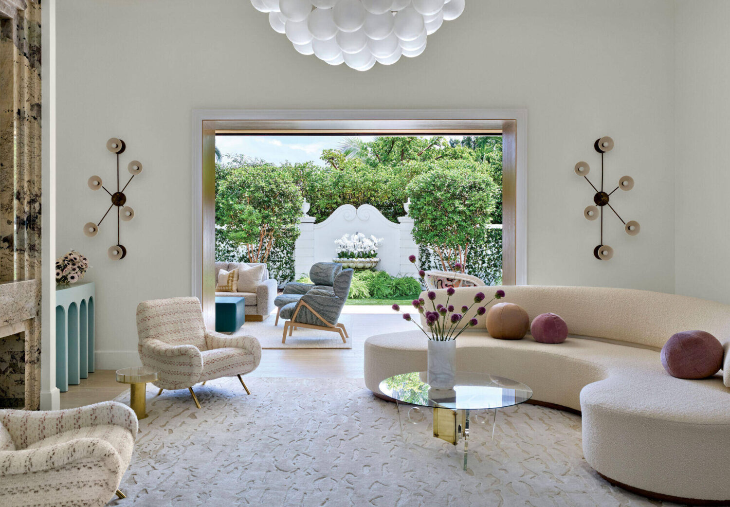 Modern living room with curved sofa, armchairs, and glass coffee table, opening onto a patio with seating and lush garden view.