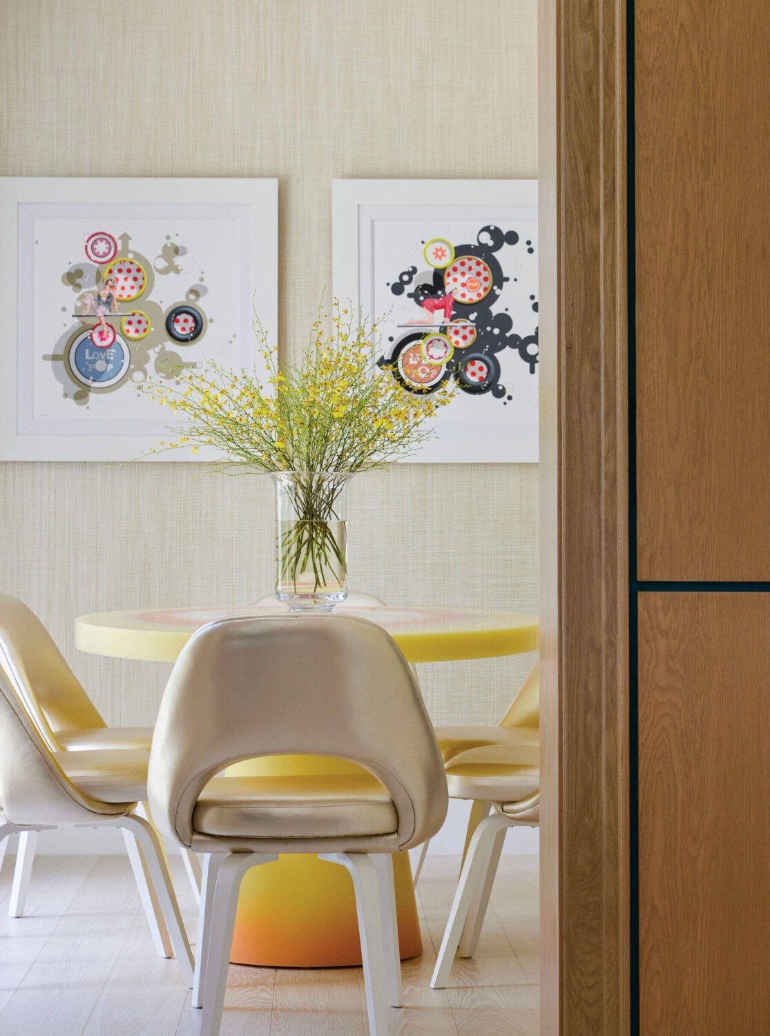 A round table with four cream chairs surrounds a vase of yellow flowers. Two abstract artworks hang on a beige wall in the background.