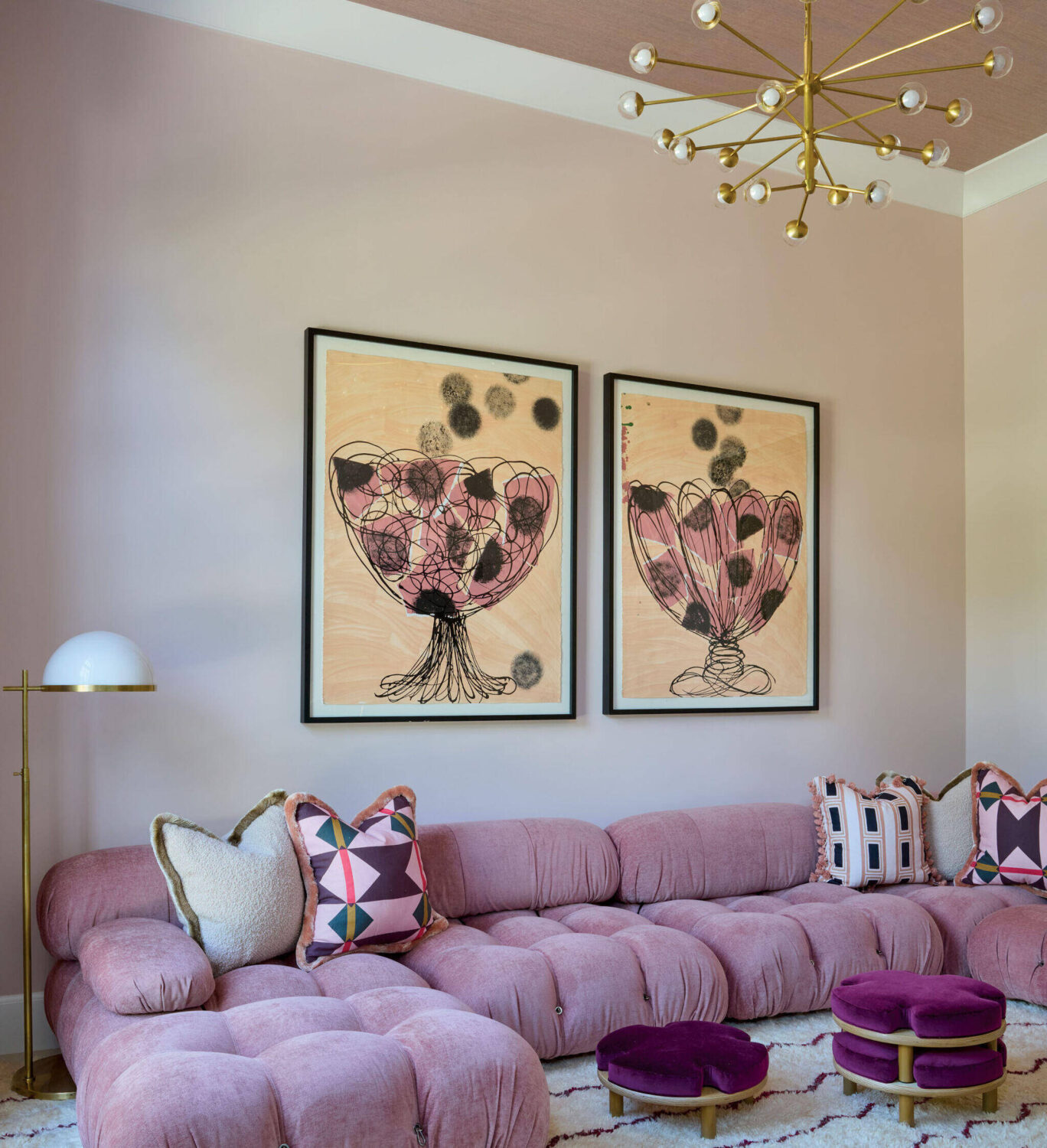 A modern living room with a pink tufted sectional sofa, geometric-patterned pillows, round purple ottomans, two abstract art pieces, and a brass sputnik chandelier.
