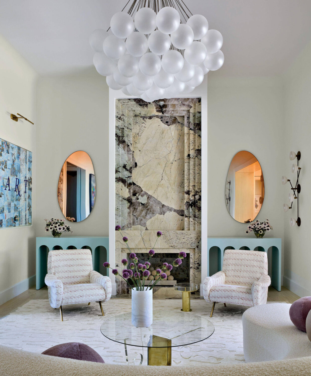Modern living room with two patterned armchairs, a marble accent wall, oval mirrors, a glass coffee table, and a cluster of white pendant lights hanging from the ceiling.