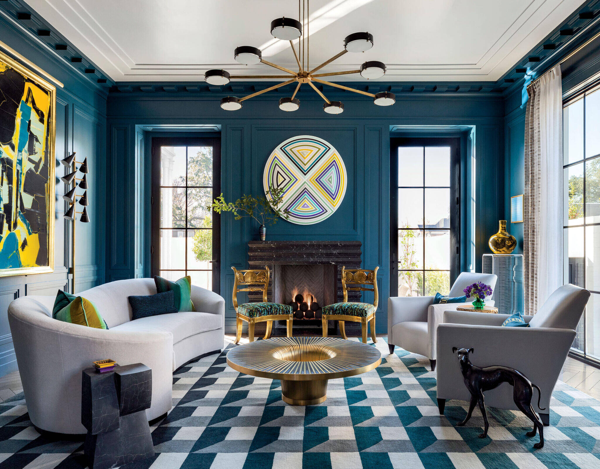 Modern living room with teal paneled walls, geometric artwork, two white sofas, a round coffee table, and a black dog sculpture, with large windows and a lit fireplace.