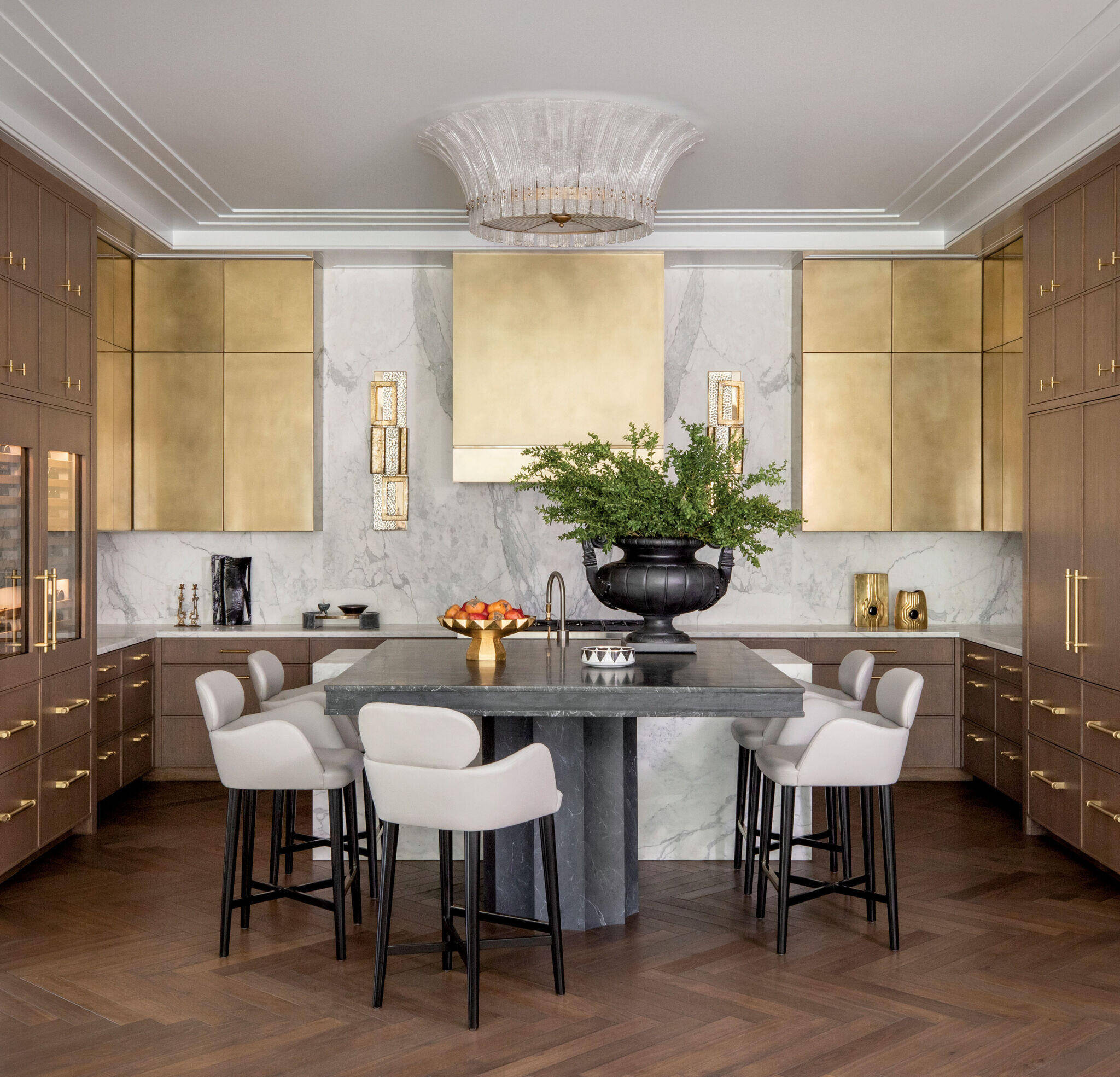 Modern kitchen with wood and gold cabinetry, marble backsplash, large island with gray countertop, white barstools, and a black vase with greenery as centerpiece.