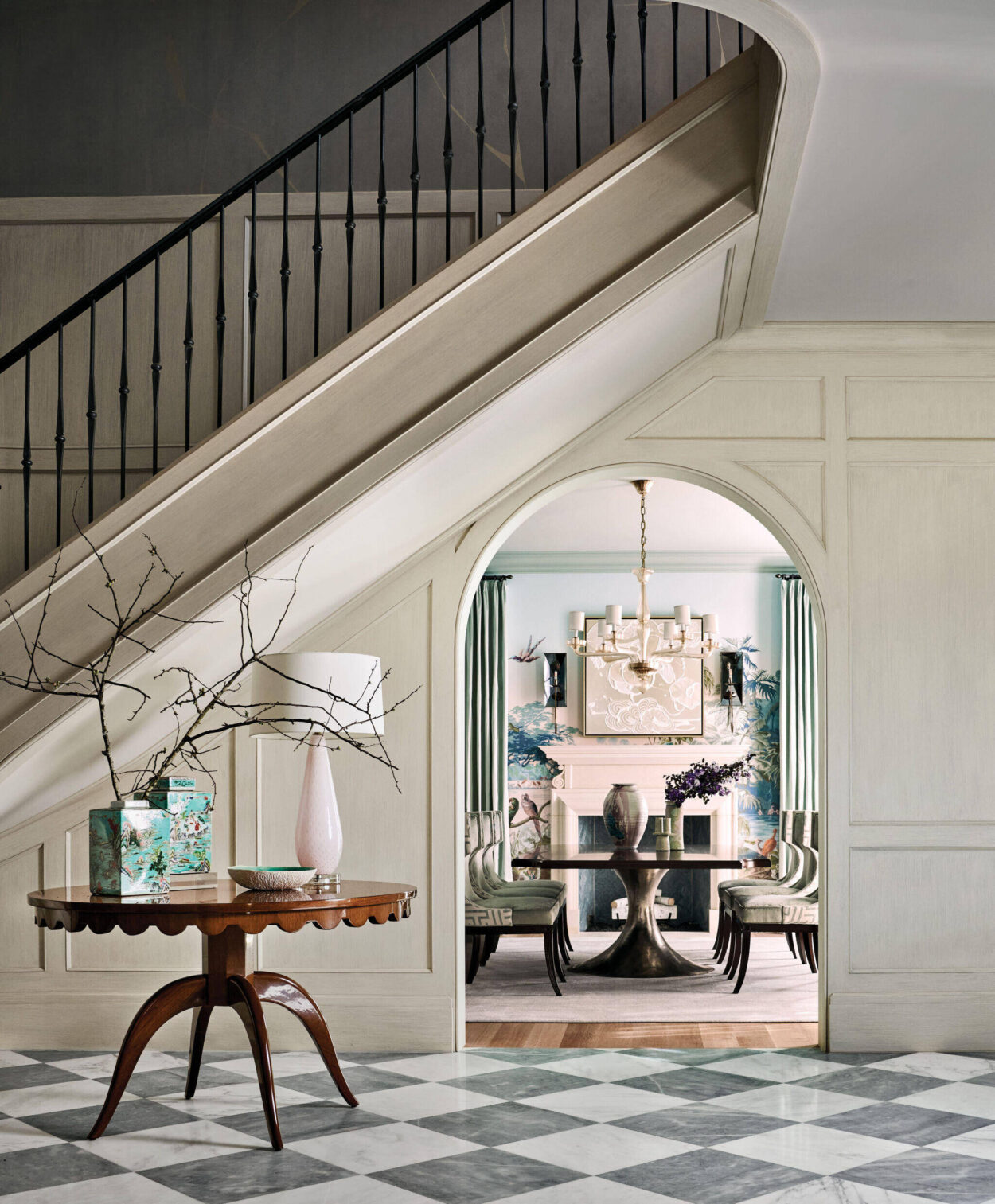 Elegant entryway with checkered floor, round table, and staircase; arched doorway leads to a stylish living room with a fireplace and contemporary decor.