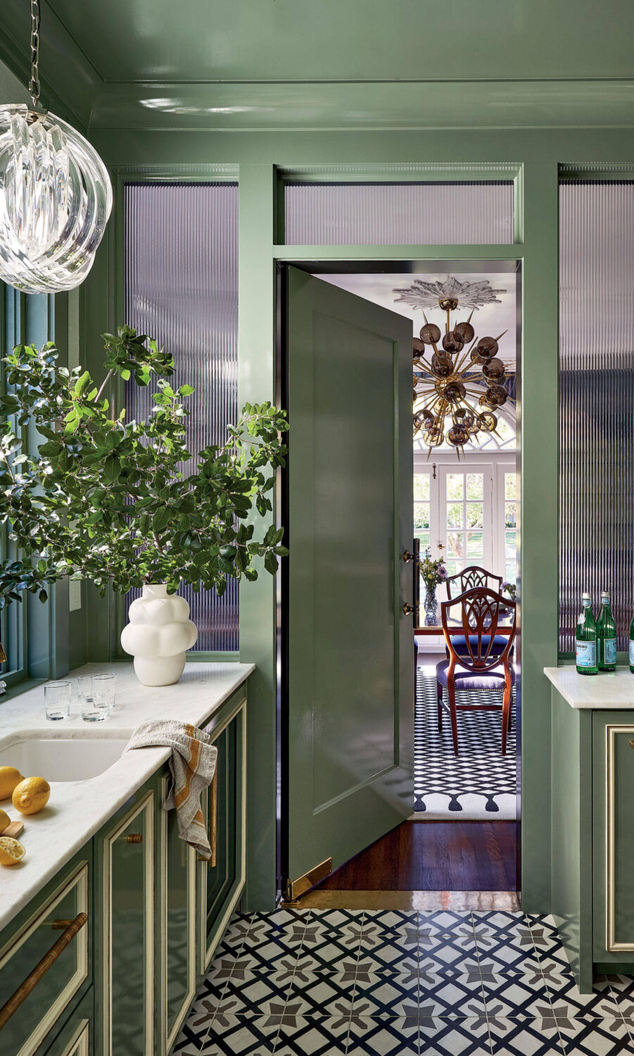 A green kitchen with a patterned tile floor, a partially open door leading to a dining room, a white vase with greenery, and pendant lighting overhead.