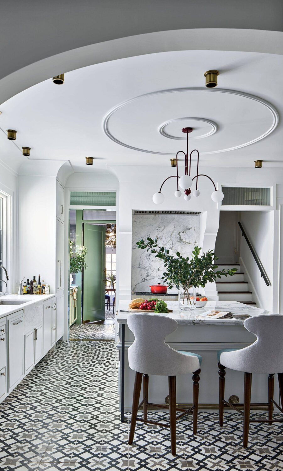 Modern kitchen with patterned tile floor, white cabinets, marble countertop, two upholstered bar stools, and a unique ceiling light fixture; stairs and a green door are visible in the background.