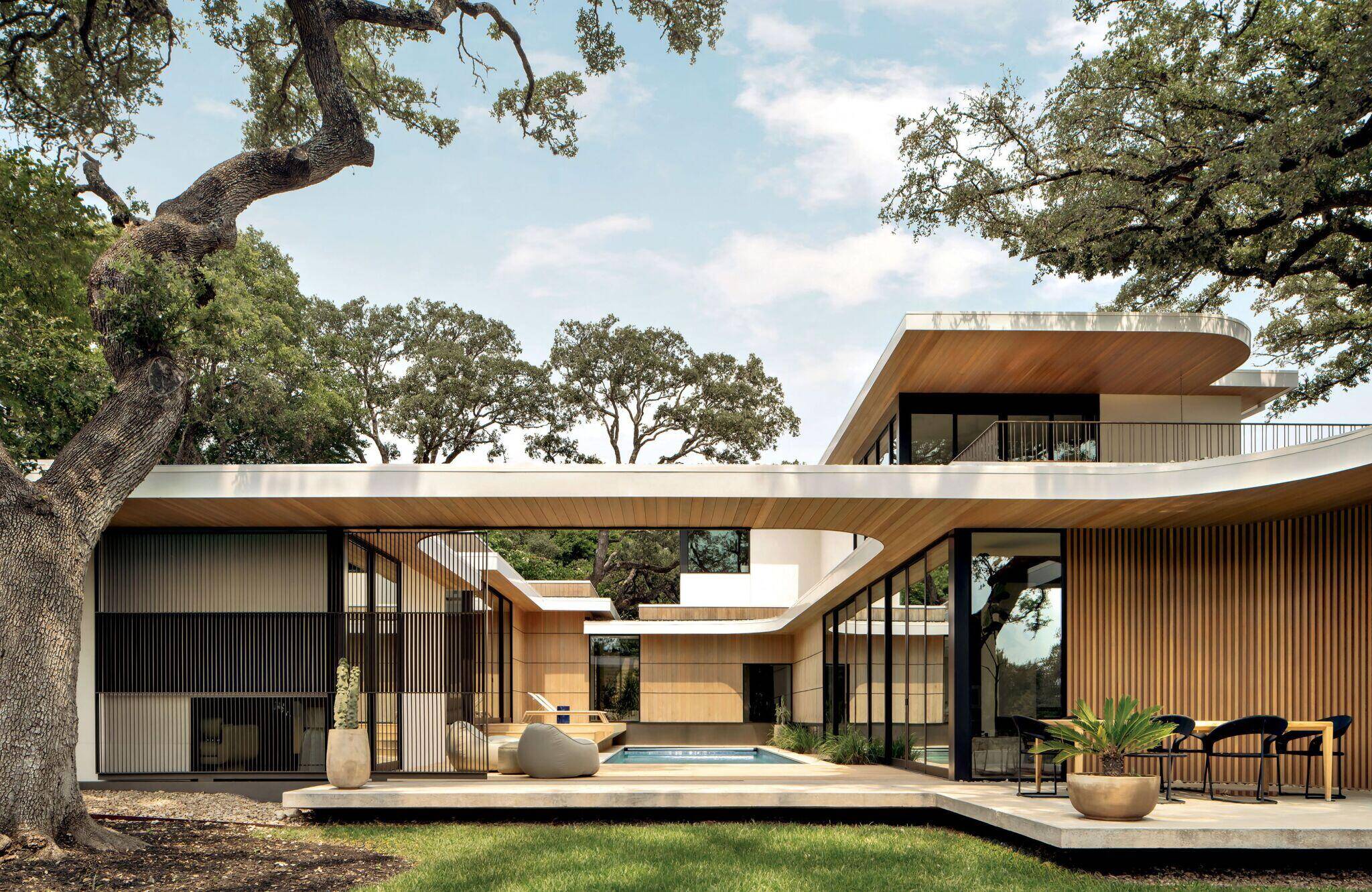 Modern house with large windows, wood paneling, and a flat roof; features an open courtyard with a small pool, outdoor seating, and surrounding trees.