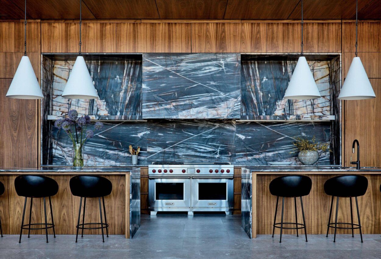 Modern kitchen with wood cabinetry, dramatic marble backsplash, stainless steel stove, black bar stools, and white pendant lights. Flowers and plants decorate the counters.