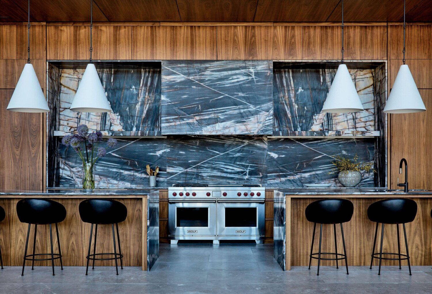 Modern kitchen with wood cabinetry, dramatic marble backsplash, stainless steel stove, black bar stools, and white pendant lights. Flowers and plants decorate the counters.