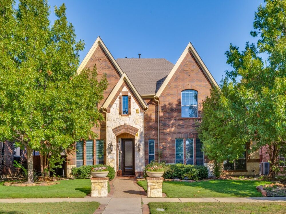 Two-story brick house with stone accents, large front windows, arched doorway, and a well-maintained lawn with trees and planters lining the walkway.