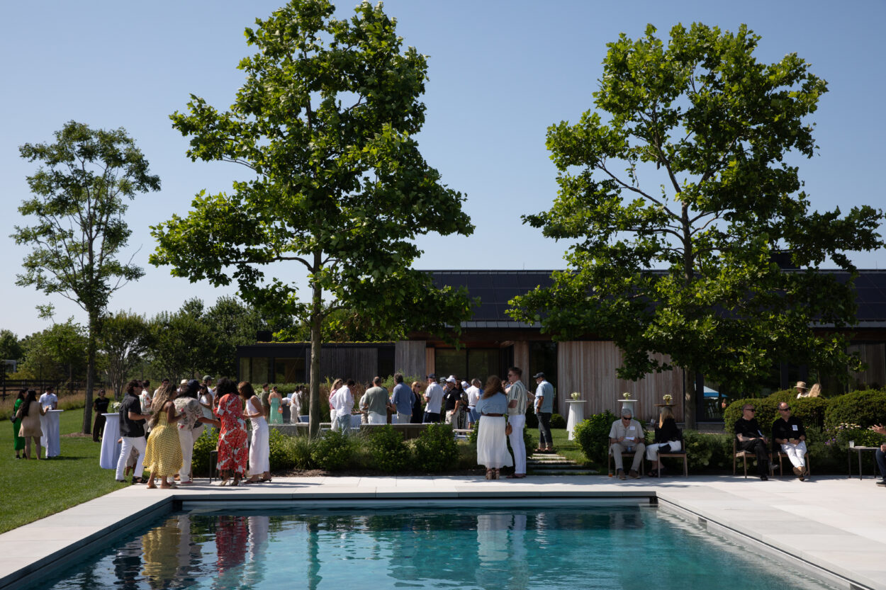 People gather beside a pool at an outdoor event on a sunny day, with trees and a modern building in the background. Some guests stand in groups while others sit in the shade.