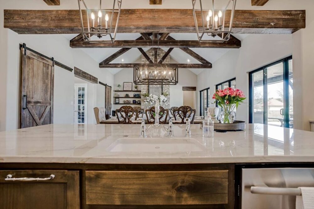 A farmhouse-style kitchen with a marble island, wooden beams, chandeliers, and sliding barn doors. Pink flowers and white flowers are on the countertop near the sink.