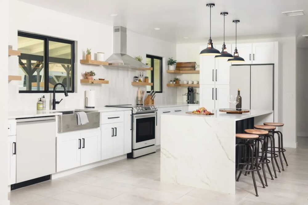 Modern kitchen with white cabinets, marble island with bar stools, stainless steel appliances, and open wooden shelves holding plants and dishes. Pendant lights hang above the island.