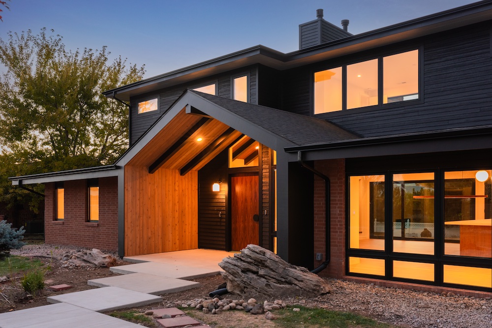 Modern two-story house with dark siding, large windows, and a wood-accented entryway, photographed at dusk with lights on inside.